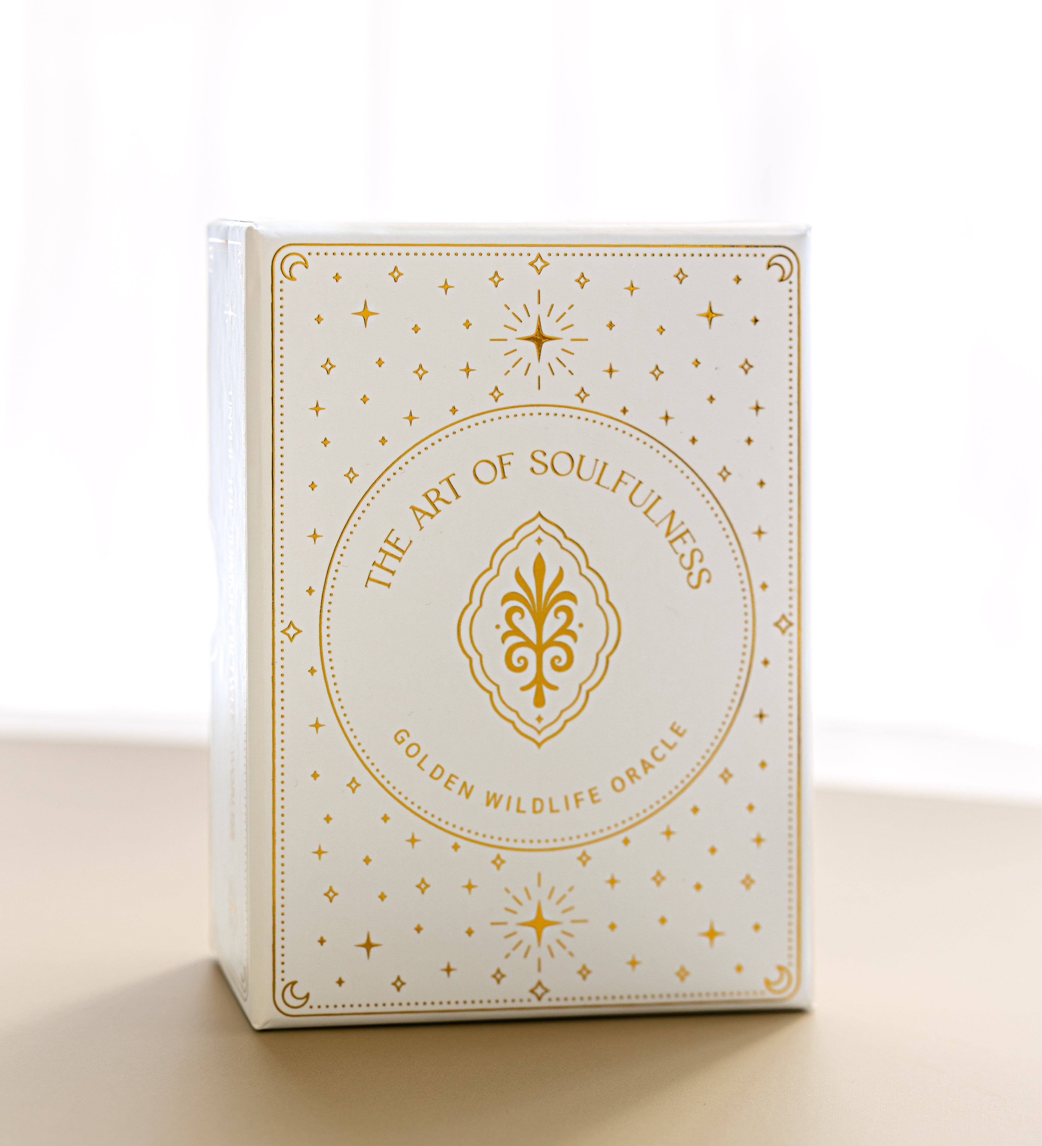 a front view of the gold foiled golden wildlife oracle box on a beige surface with a sheer curtain blurred in the background
