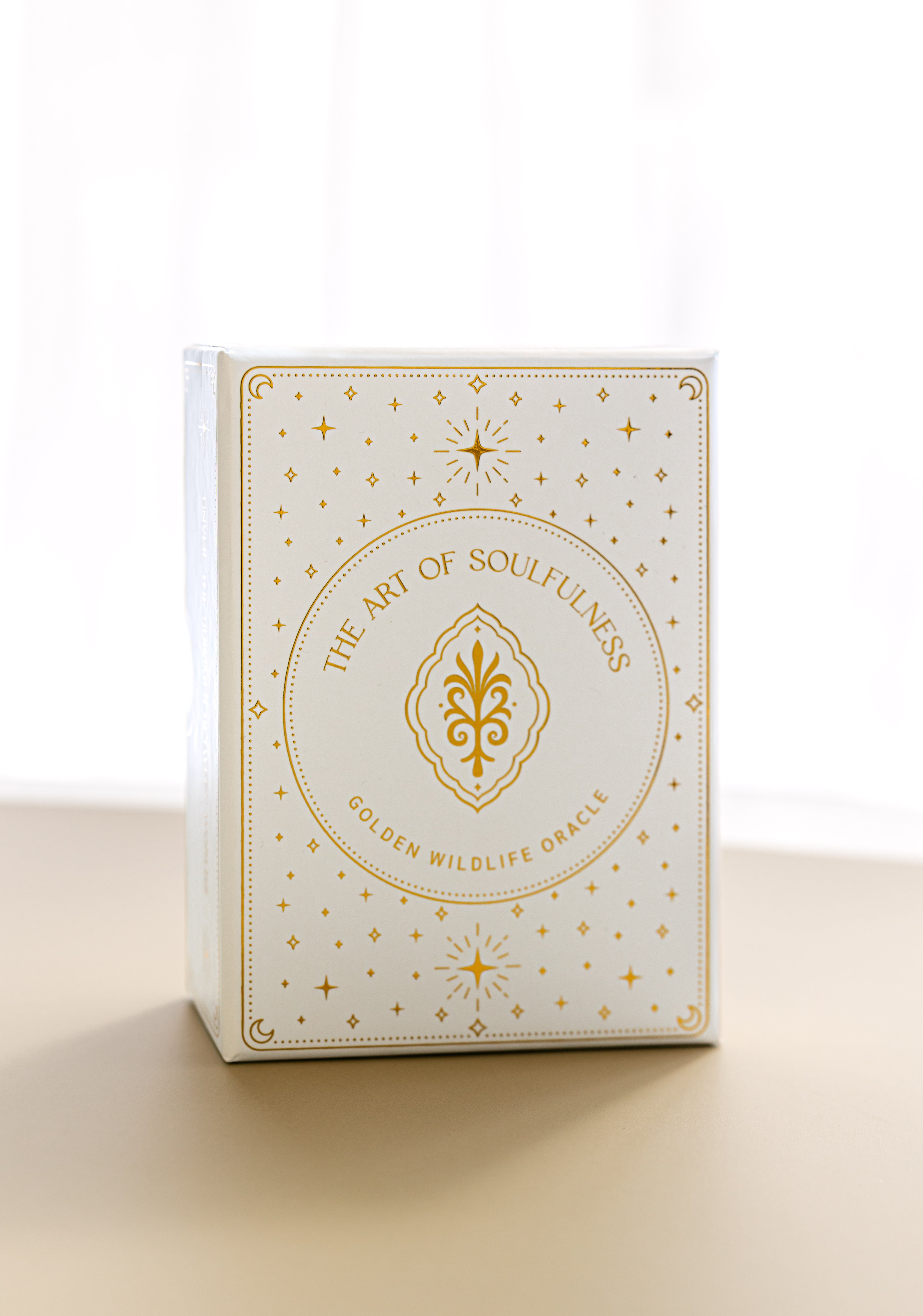 a front view of the gold foiled golden wildlife oracle box on a beige surface with a sheer curtain blurred in the background