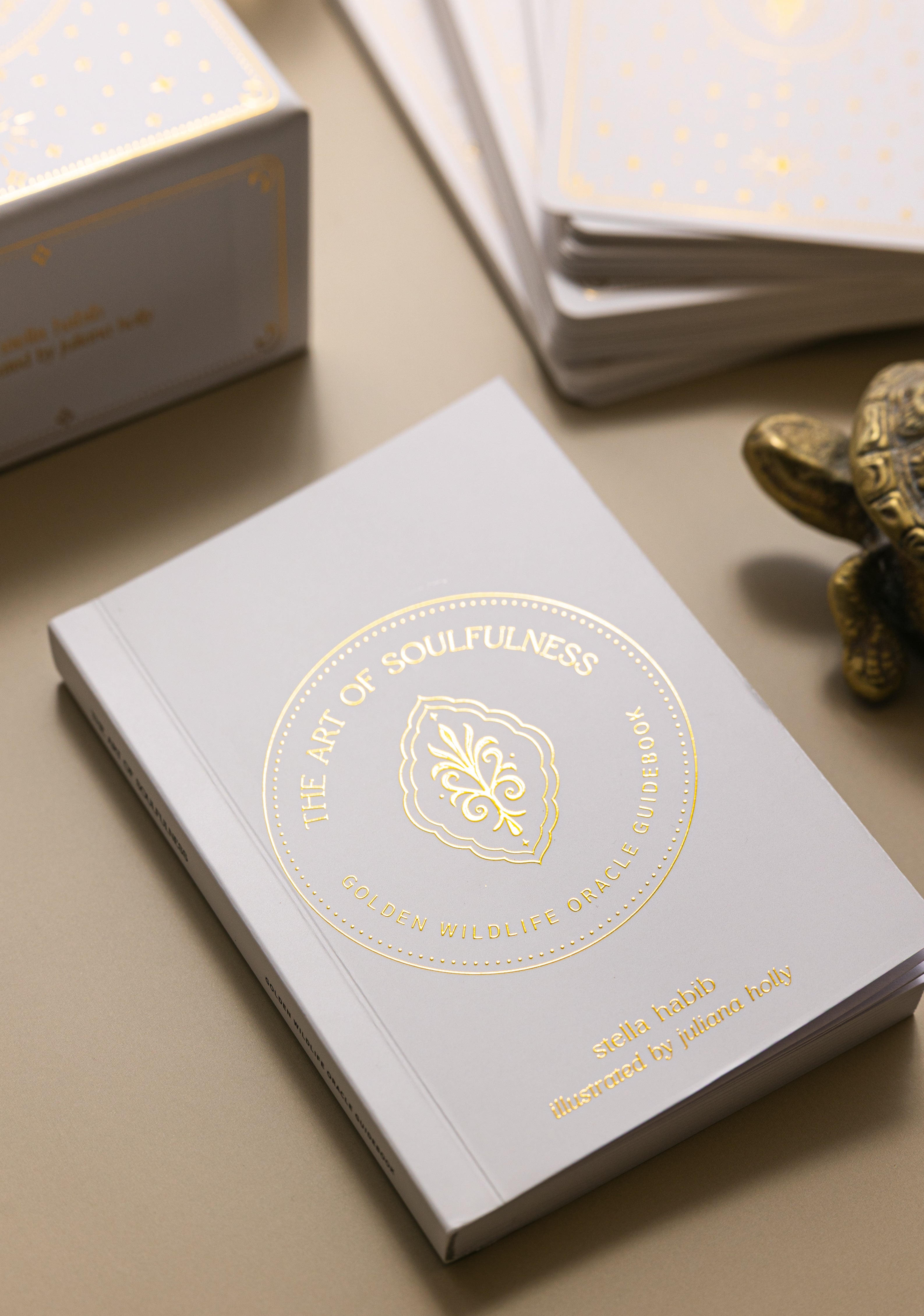 glimmering gold on the cover of the golden wildlife oracle guidebook with the box and cards and the head of a brass turtle in the background.