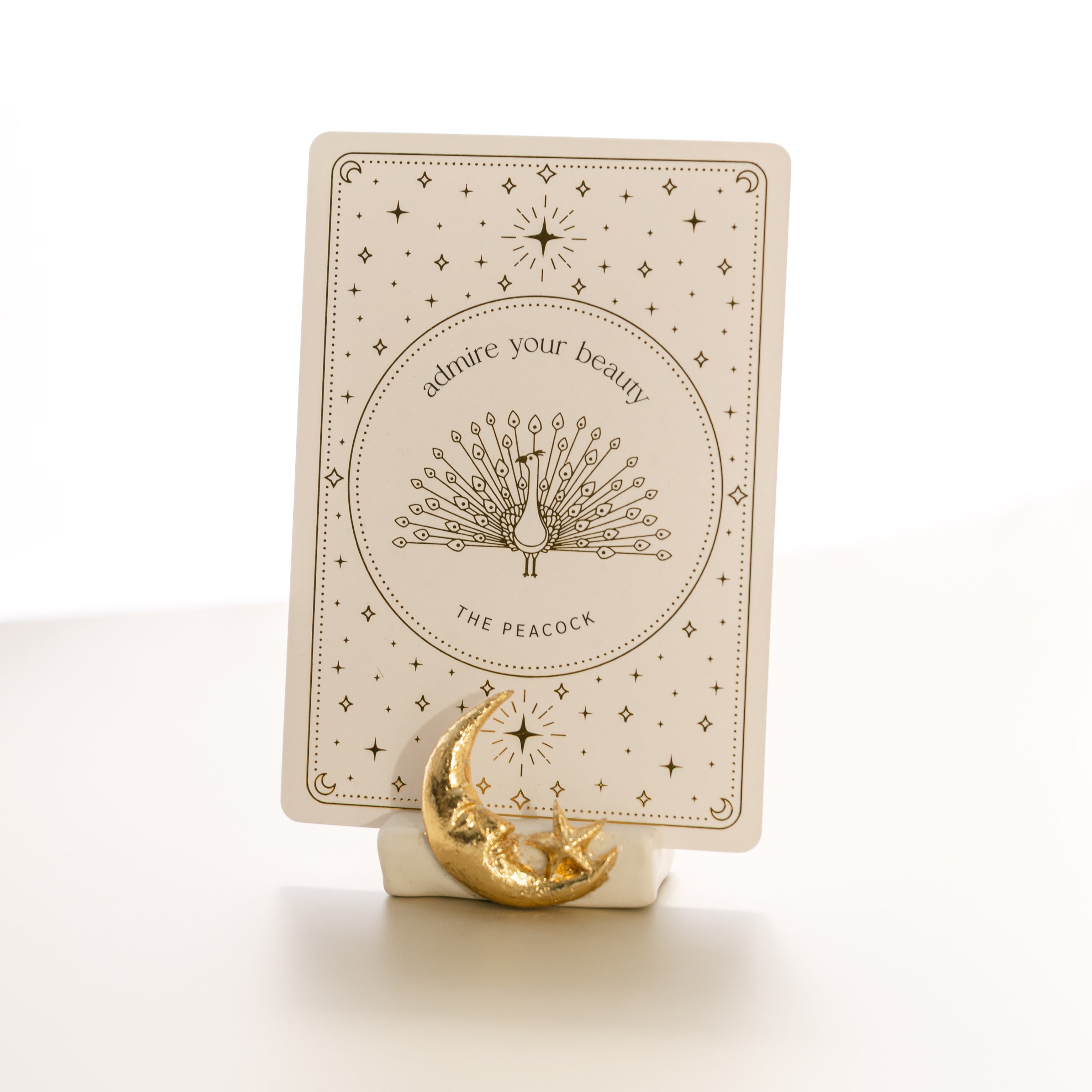 oracle card with a gold foil design that says 'admire your beauty' and features a peacock, held by a matte white card stand decorated with a sculpted gold foil moon and star.