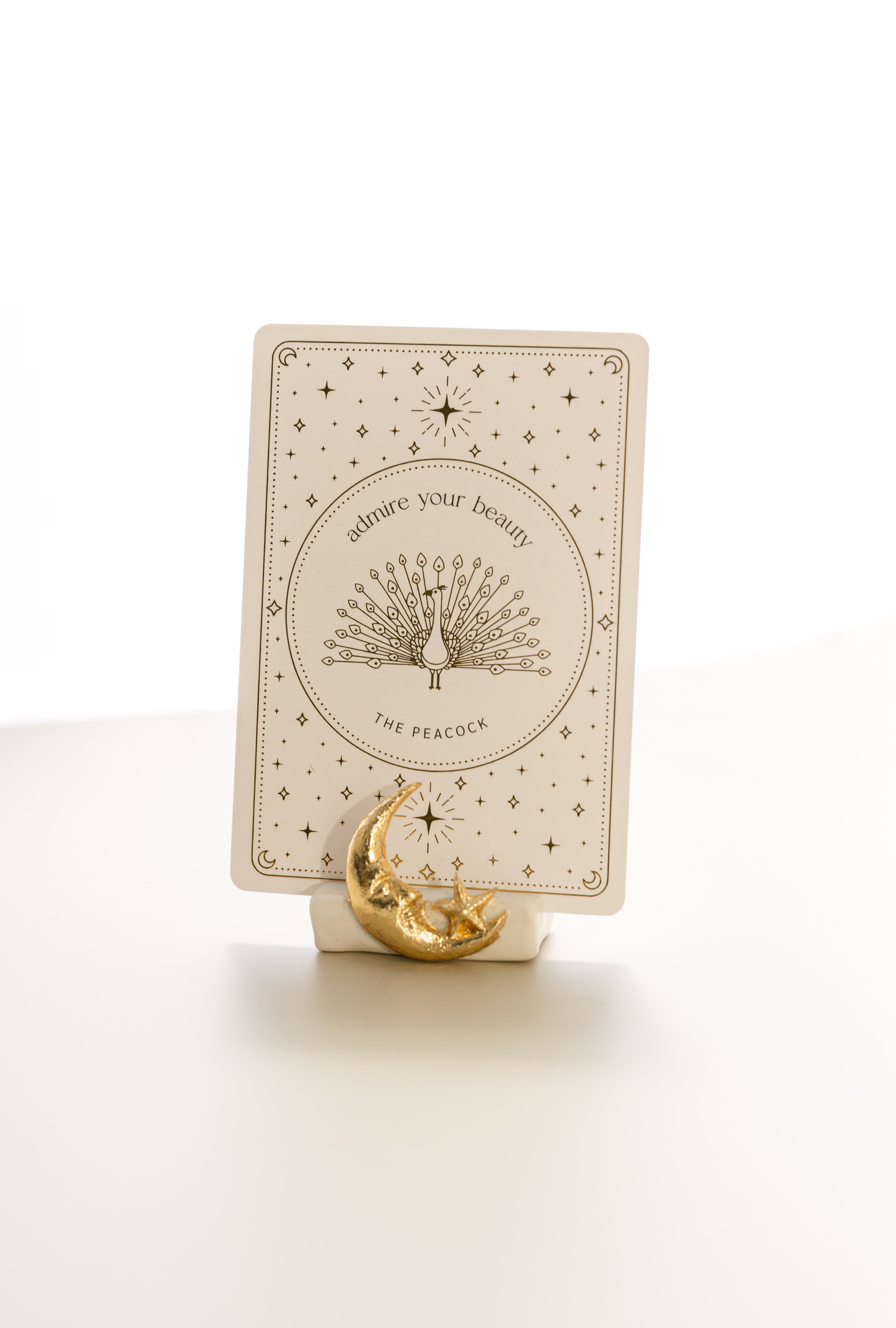 oracle card with a gold foil design that says 'admire your beauty' and features a peacock, held by a matte white card stand decorated with a sculpted gold foil moon and star.