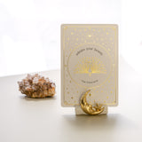 a white matte ceramic oracle card holder with a sculpted moon and star, decorated in gold foil, displaying the peacock card from the golden wildlife oracle with a red aragonite crystal in the background
