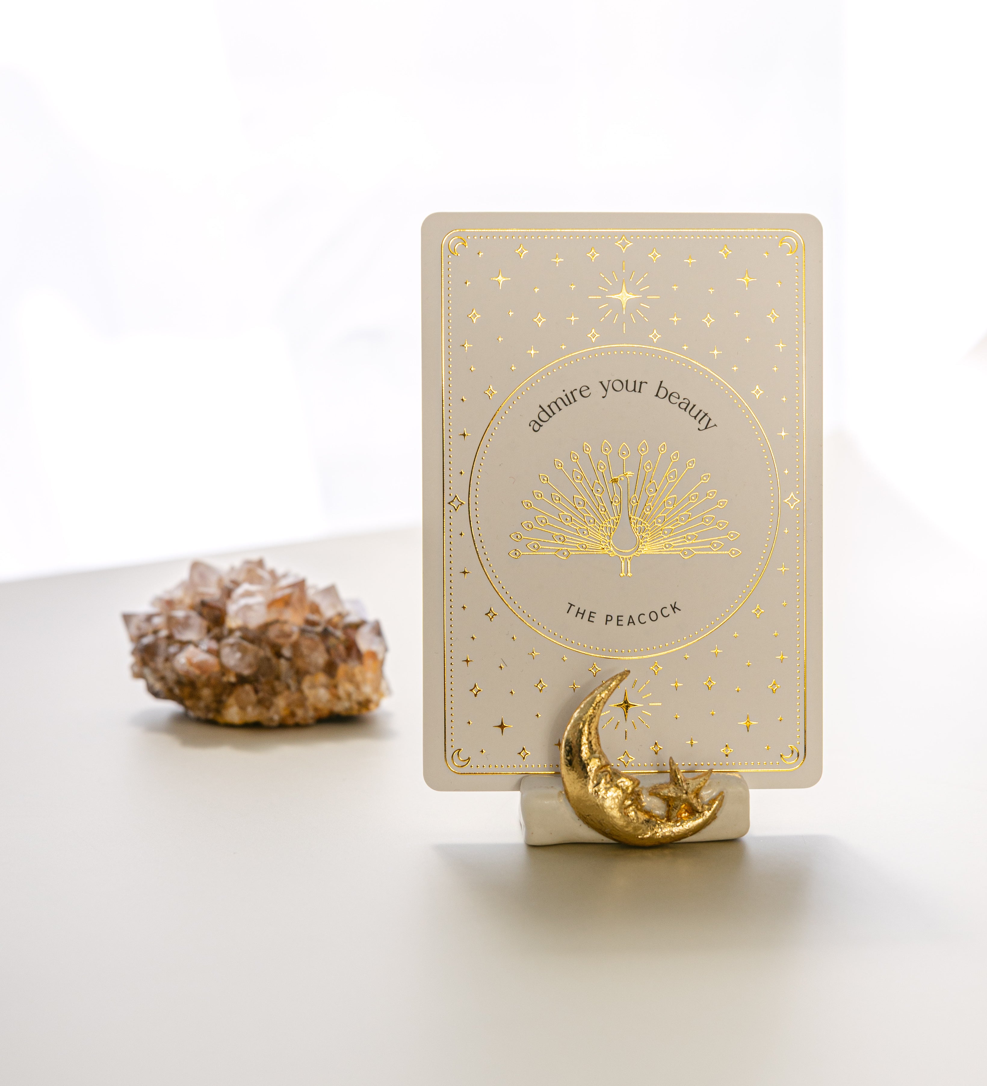 a white matte ceramic oracle card holder with a sculpted moon and star, decorated in gold foil, displaying the peacock card from the golden wildlife oracle with a red aragonite crystal in the background