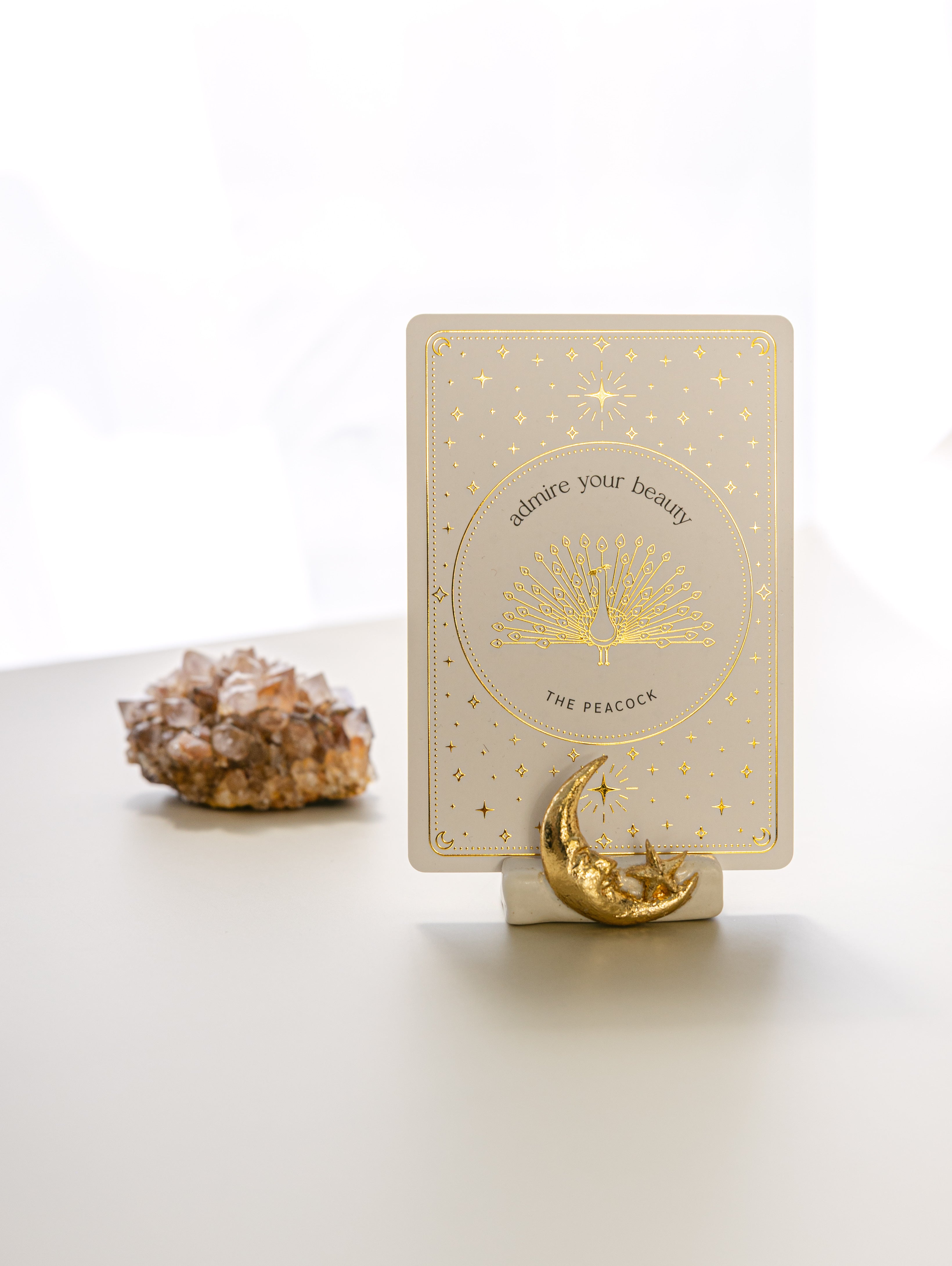 a white matte ceramic oracle card holder with a sculpted moon and star, decorated in gold foil, displaying the peacock card from the golden wildlife oracle with a red aragonite crystal in the background