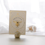 a pearlescent ceramic orb oracle card stand holding the bee card from teh golden wildlife oracle with a white quartz crystal and sheer curtain in the background.