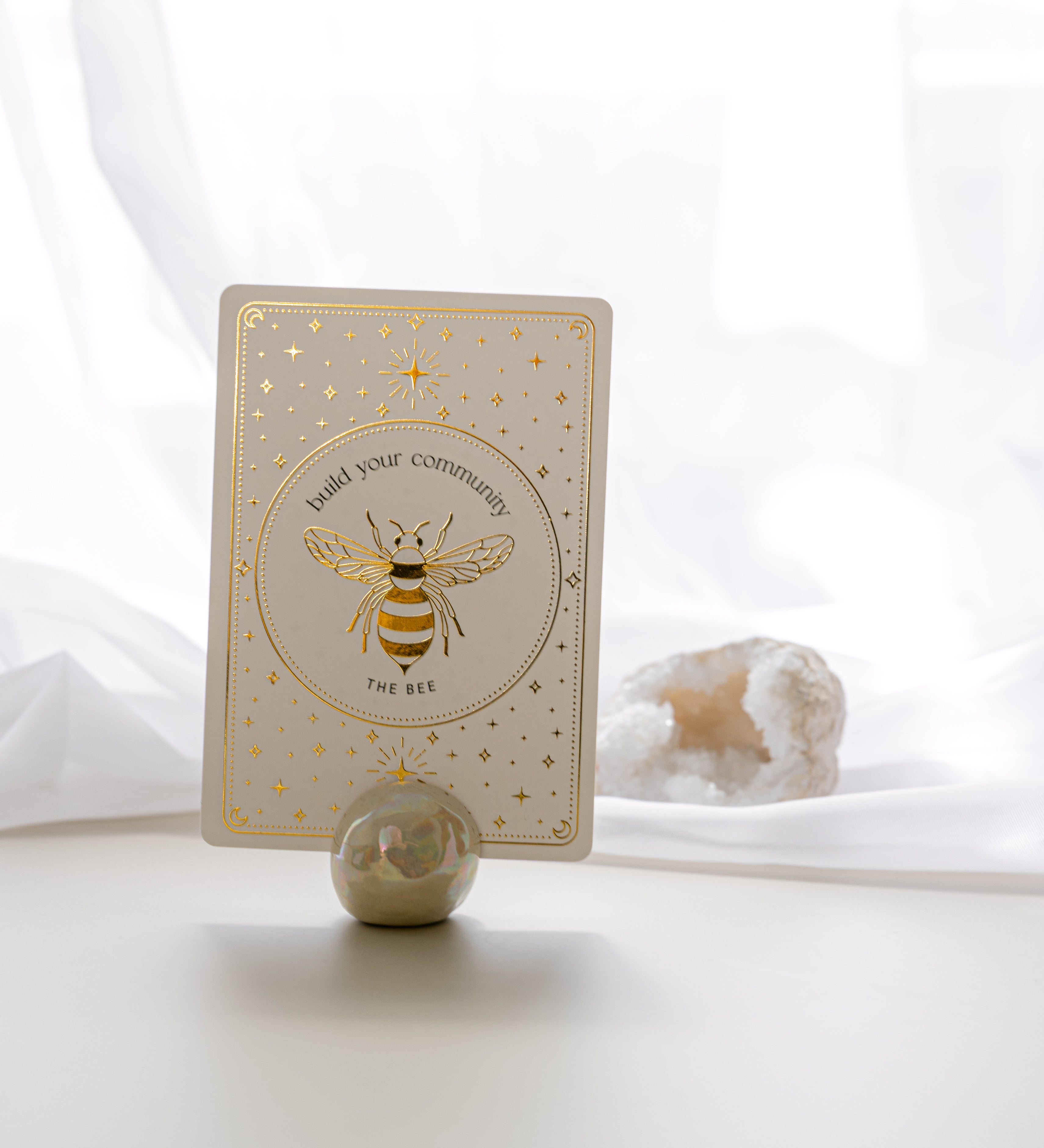 a pearlescent ceramic orb oracle card stand holding the bee card from teh golden wildlife oracle with a white quartz crystal and sheer curtain in the background.