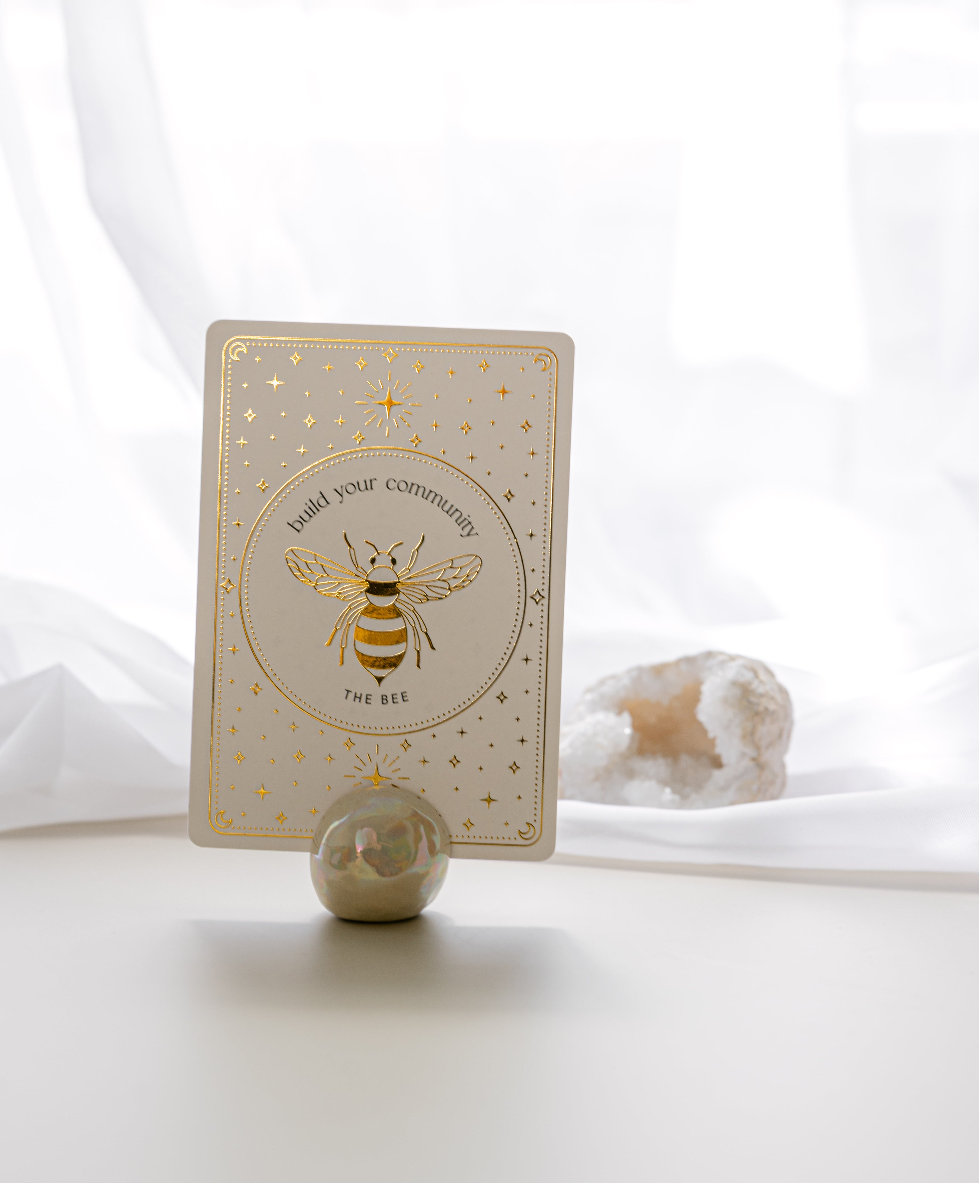 a pearlescent ceramic orb oracle card stand holding the bee card from teh golden wildlife oracle with a white quartz crystal and sheer curtain in the background.