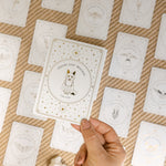 a hand holding a face-up golden wildlife oracle card with the message 'choose your direction' and an illustration of a fox, with a grid of many other face-up cards, a brass turtle and a white aragonite crystal blurred in the background