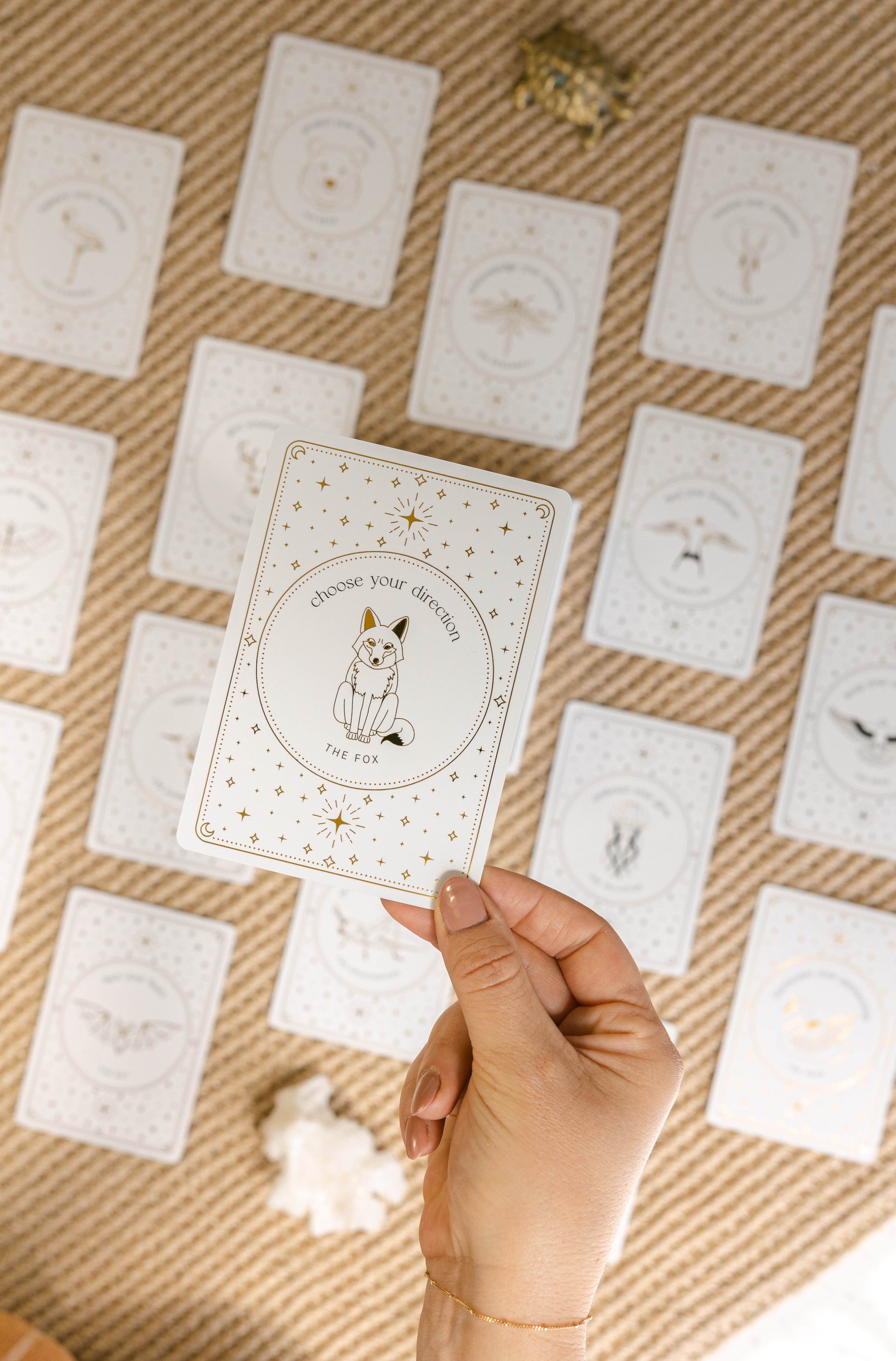 a hand holding a face-up golden wildlife oracle card with the message 'choose your direction' and an illustration of a fox, with a grid of many other face-up cards, a brass turtle and a white aragonite crystal blurred in the background