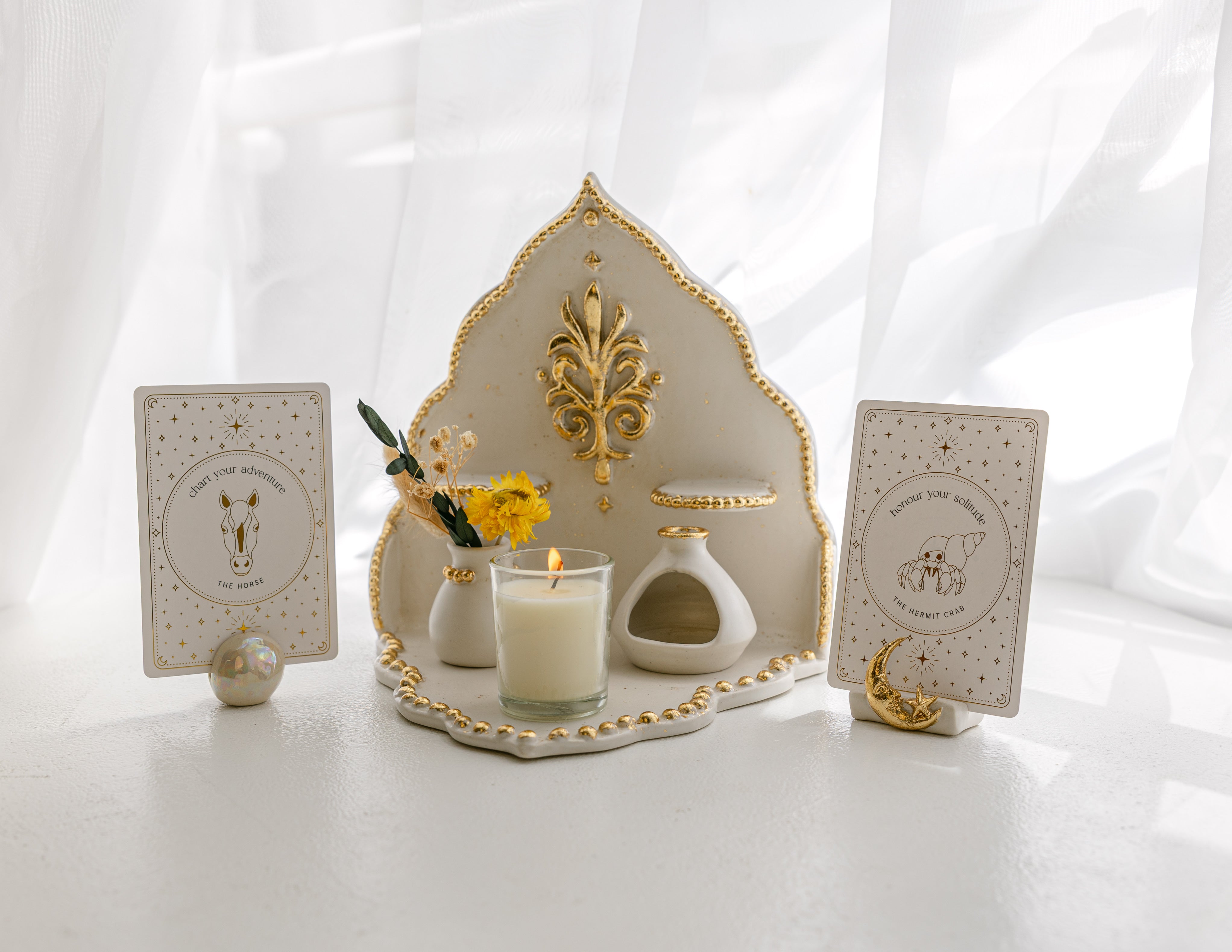 a handmade, ceramic ritual shelf featuring a sculpted art of soulfulness logo in gold, burning candle and two ceramic card stands holding golden wildlife oracle cards - the orb stand holding the 'chart your adventure' message with the illustration of a horse, and the moon stand holding the 'honour your solitude' message with the illustration of a hermit crab.