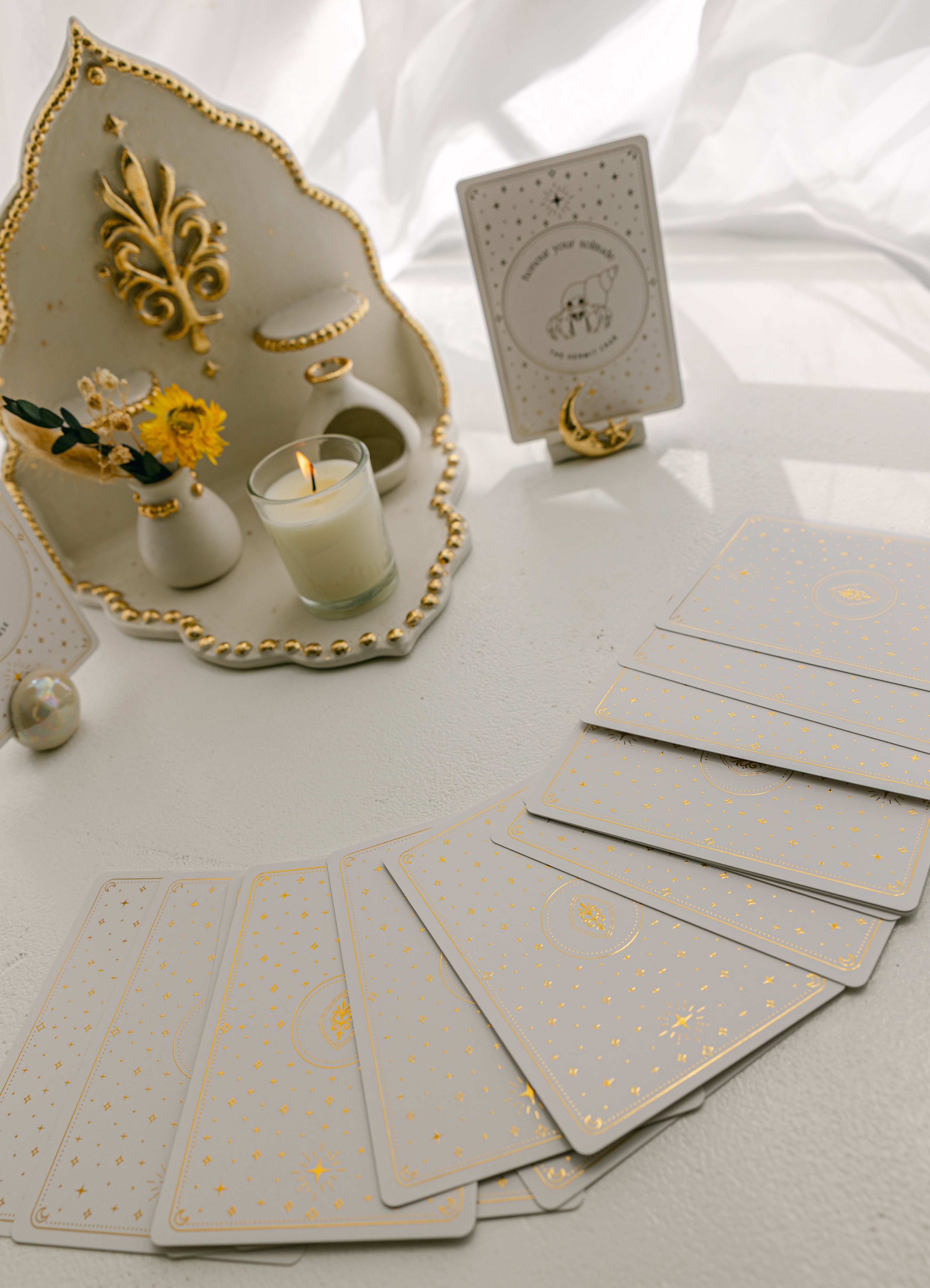 a white matte ceramic oracle card holder with a sculpted moon and star, decorated in gold foil, displaying the hermit crab card from the golden wildlife oracle with a burning candle, ritual shelf, ceramic orb card stand and face down cards from the deck nearby.