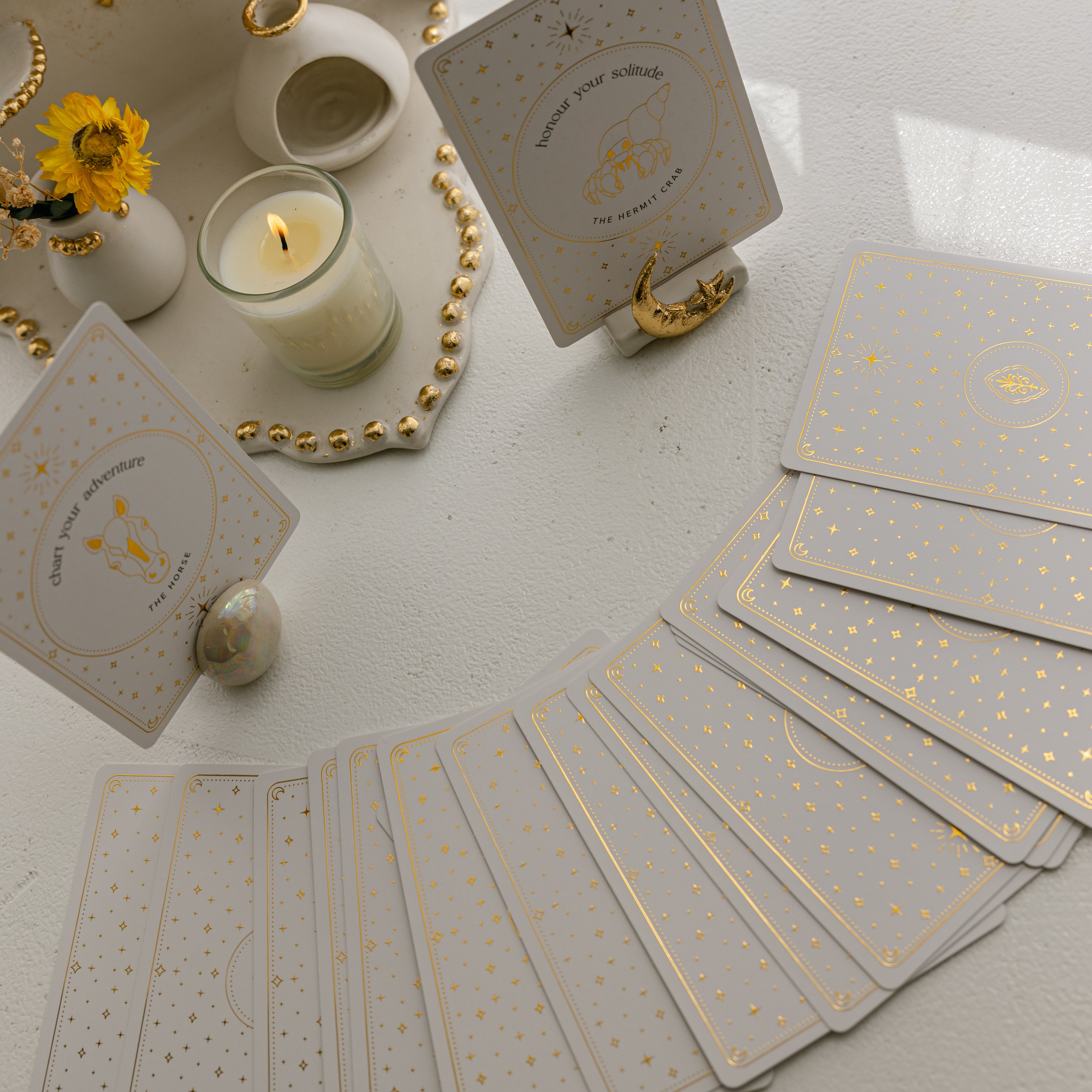 an overhead perspective of a face down arc of golden wildlife oracle cards near two featured cards, in ceramic orb and celestial ceramic card stands, alongside a ceramic ritual shelf with a burning candle.