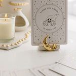 a white matte ceramic oracle card holder with a sculpted moon and star, decorated in gold foil, displaying the hermit crab card from the golden wildlife oracle with a burning candle, ritual shelf and face down cards from the deck nearby.