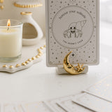 a white matte ceramic oracle card holder with a sculpted moon and star, decorated in gold foil, displaying the hermit crab card from the golden wildlife oracle with a burning candle, ritual shelf and face down cards from the deck nearby.