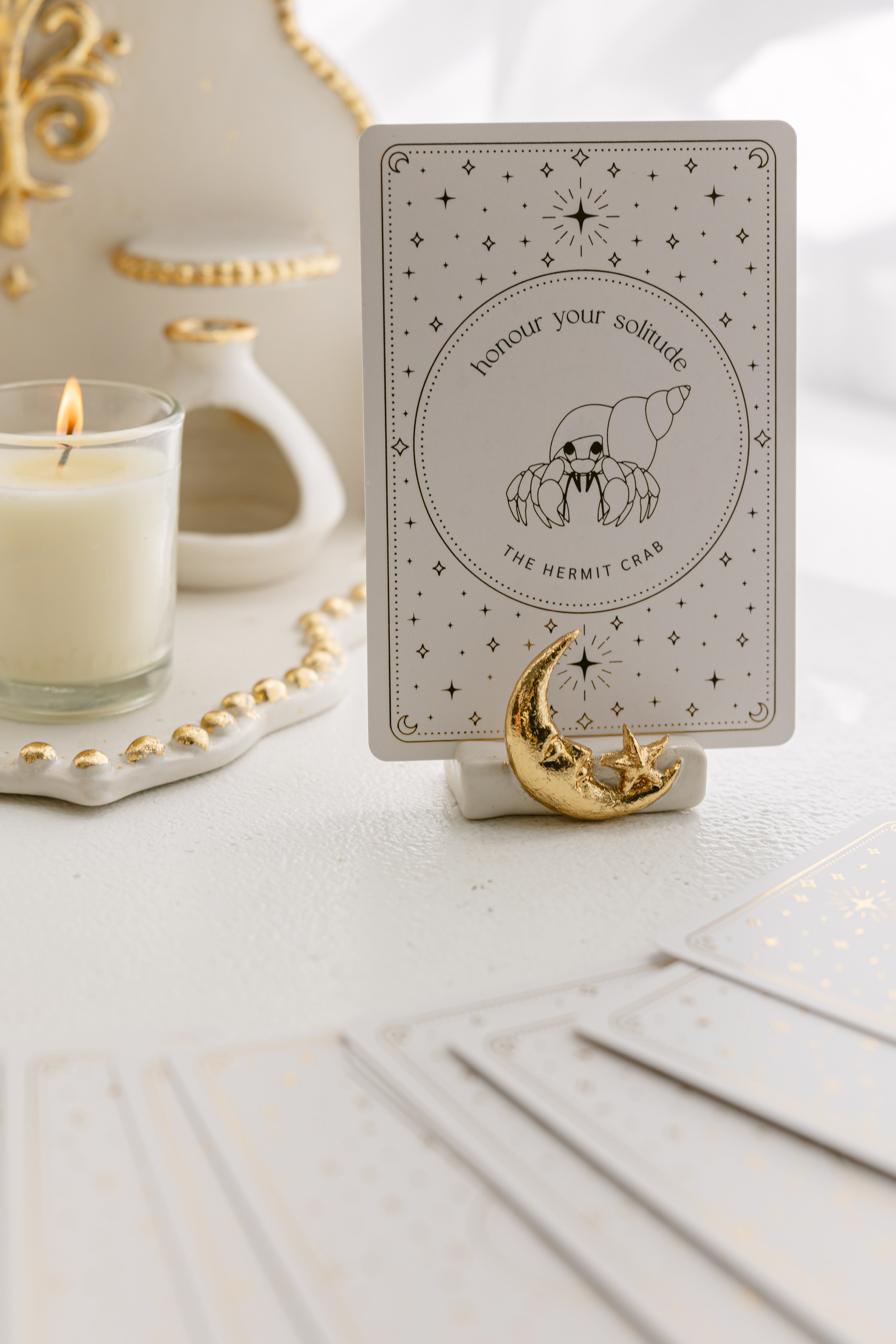 a white matte ceramic oracle card holder with a sculpted moon and star, decorated in gold foil, displaying the hermit crab card from the golden wildlife oracle with a burning candle, ritual shelf and face down cards from the deck nearby.