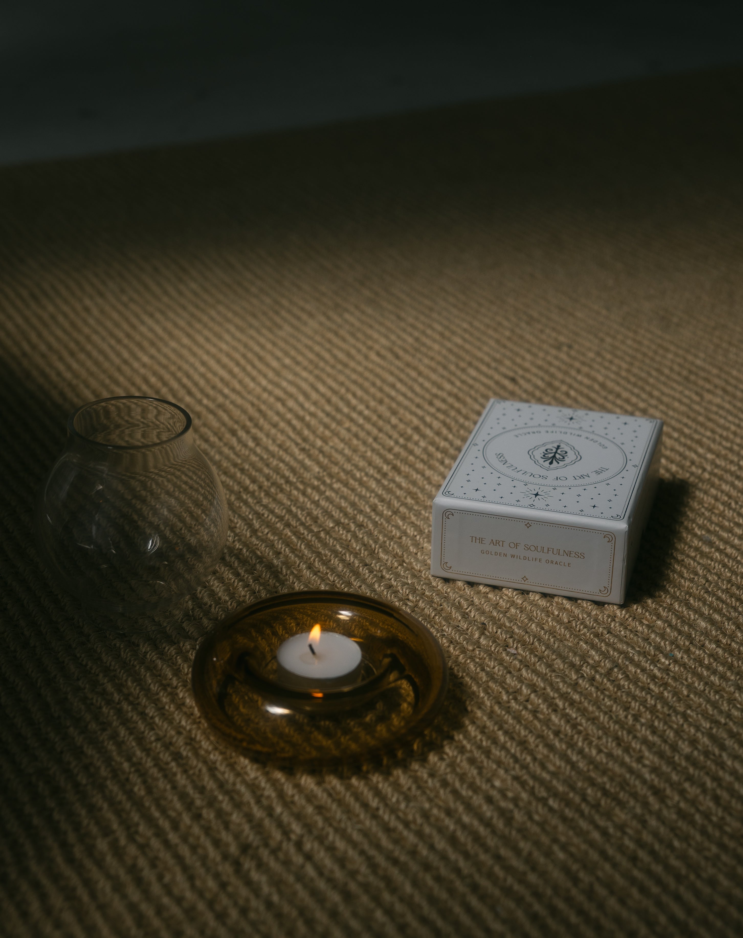 a burning tea light candle in a glass holder with the golden wildlife oracle boxed set nearby, on a braided carpet.