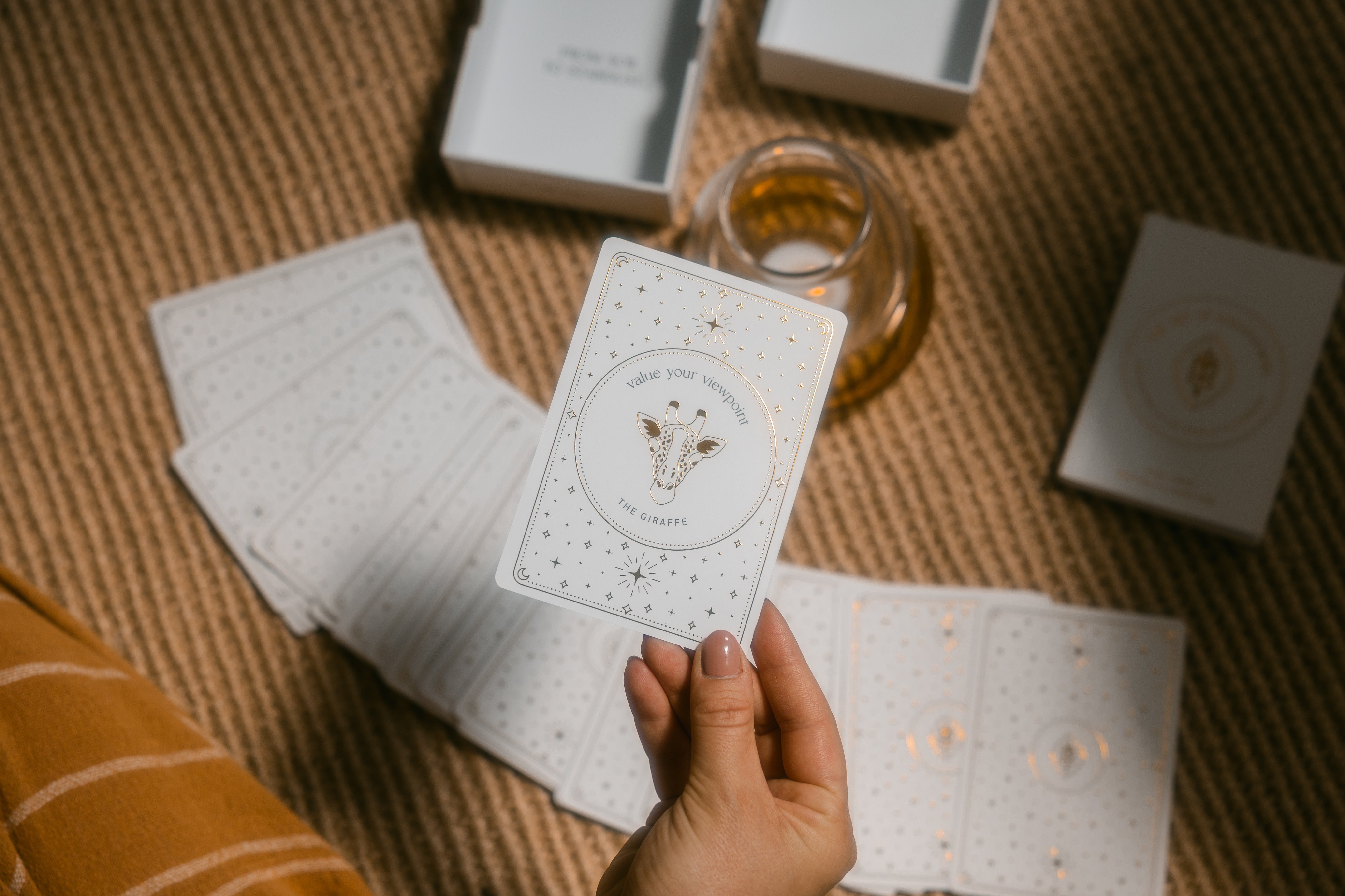 hand holding a golden wildlife oracle card over the spread out, face down deck on a braided carpet with the box, guidebook and a candle burning in the background.