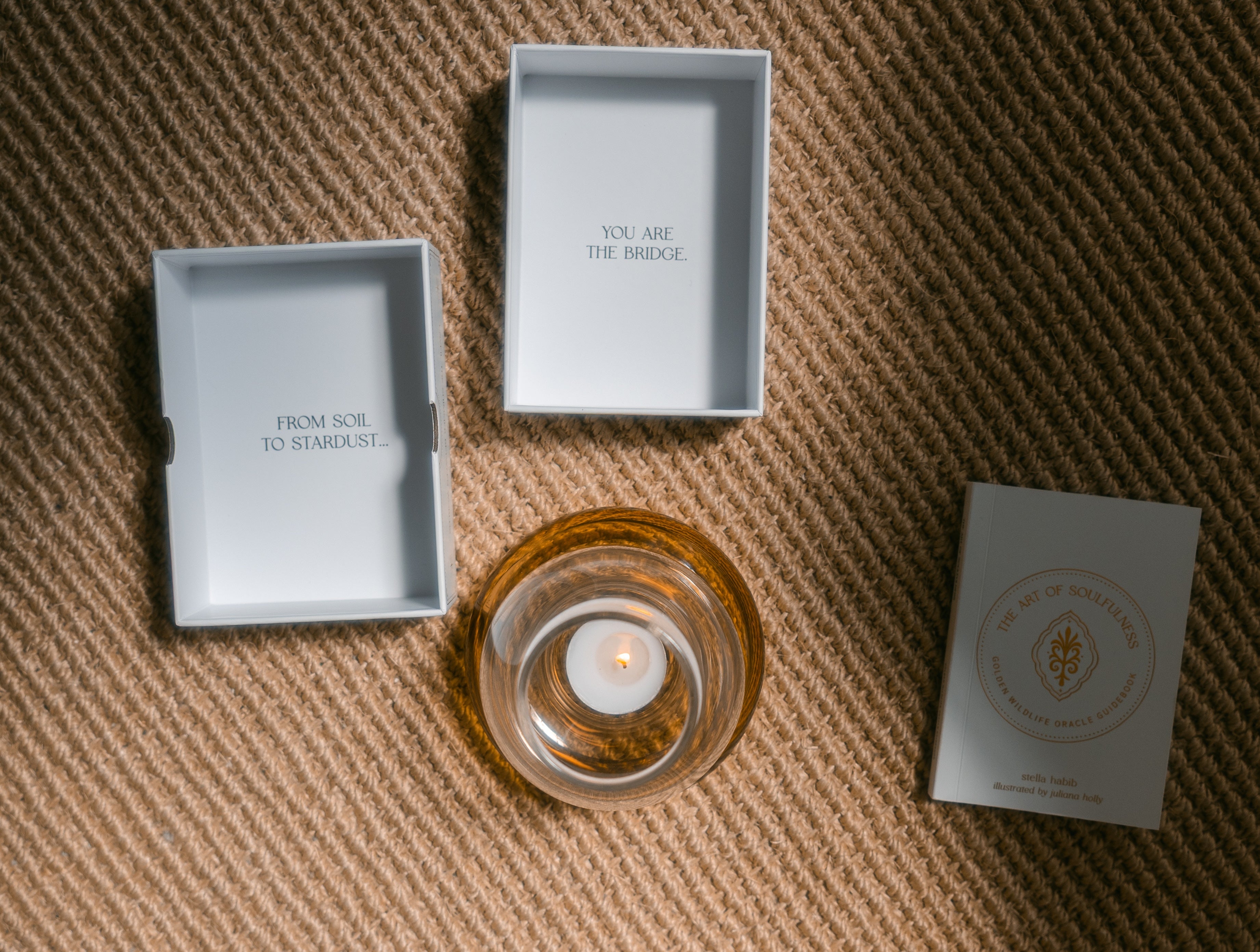 a burning tea light candle in a glass holder near the open lid and tray of the golden wildlife oracle box, which say 'from soil to stardust...' 'you are the bridge.' and the guidebook nearby, on a braided carpet.