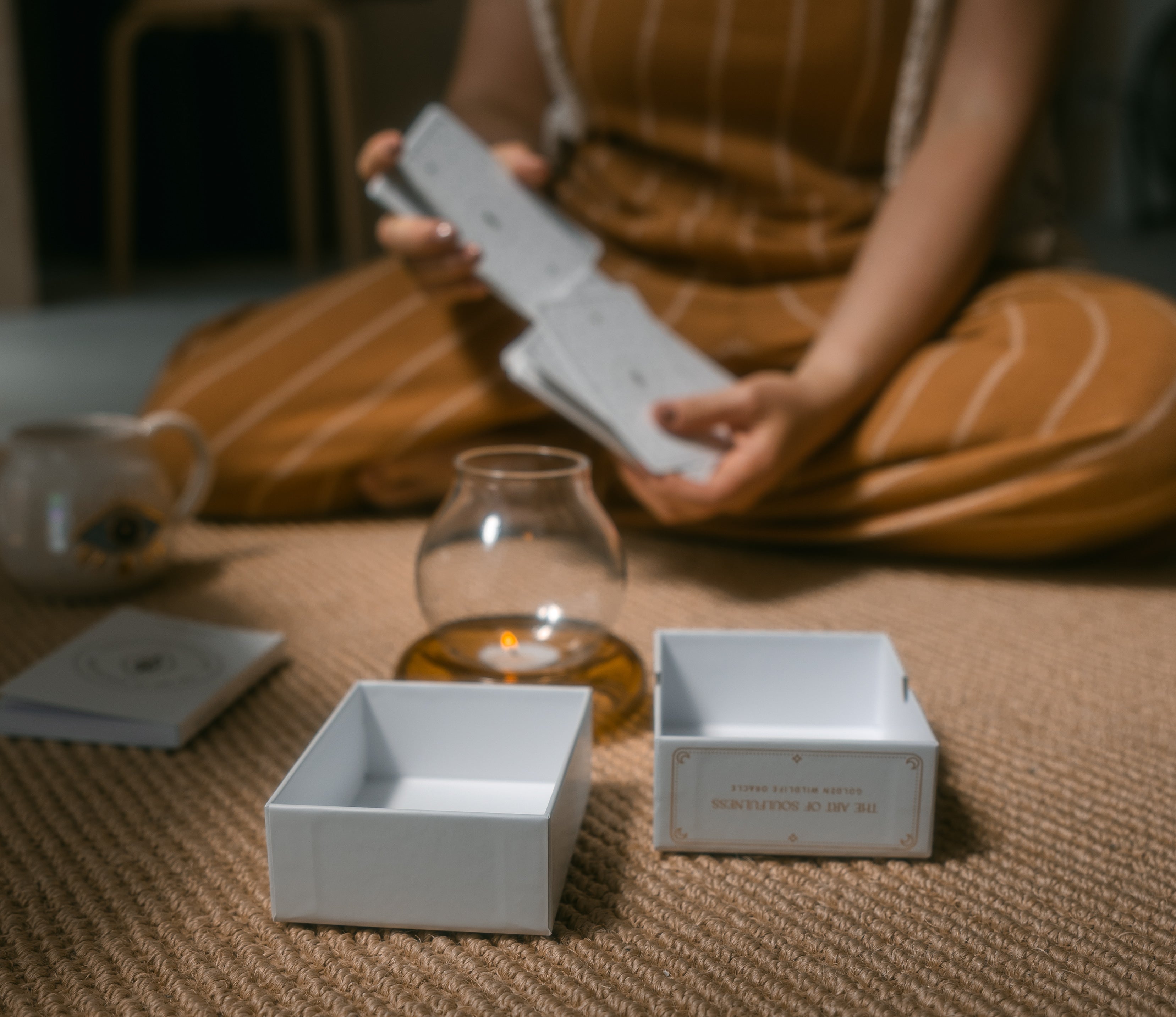 person sitting on a braided carpet while shuffling golden wildlife oracle cards, with the box, guidebook, candle and mug nearby.