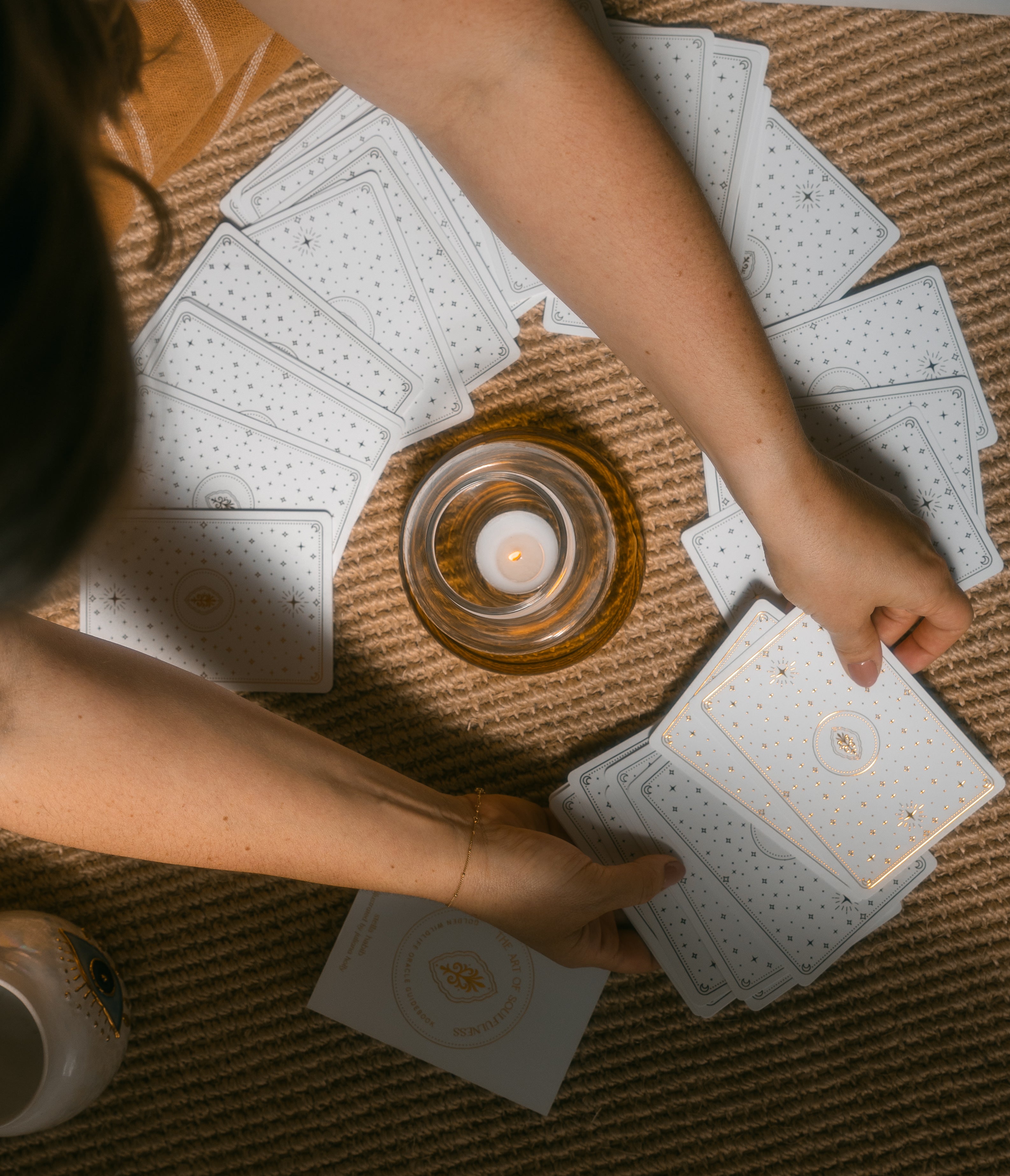 an overhead shot of a person arranging face down golden wildlife oracle cards on a braided carpet with a candle and mug nearby.