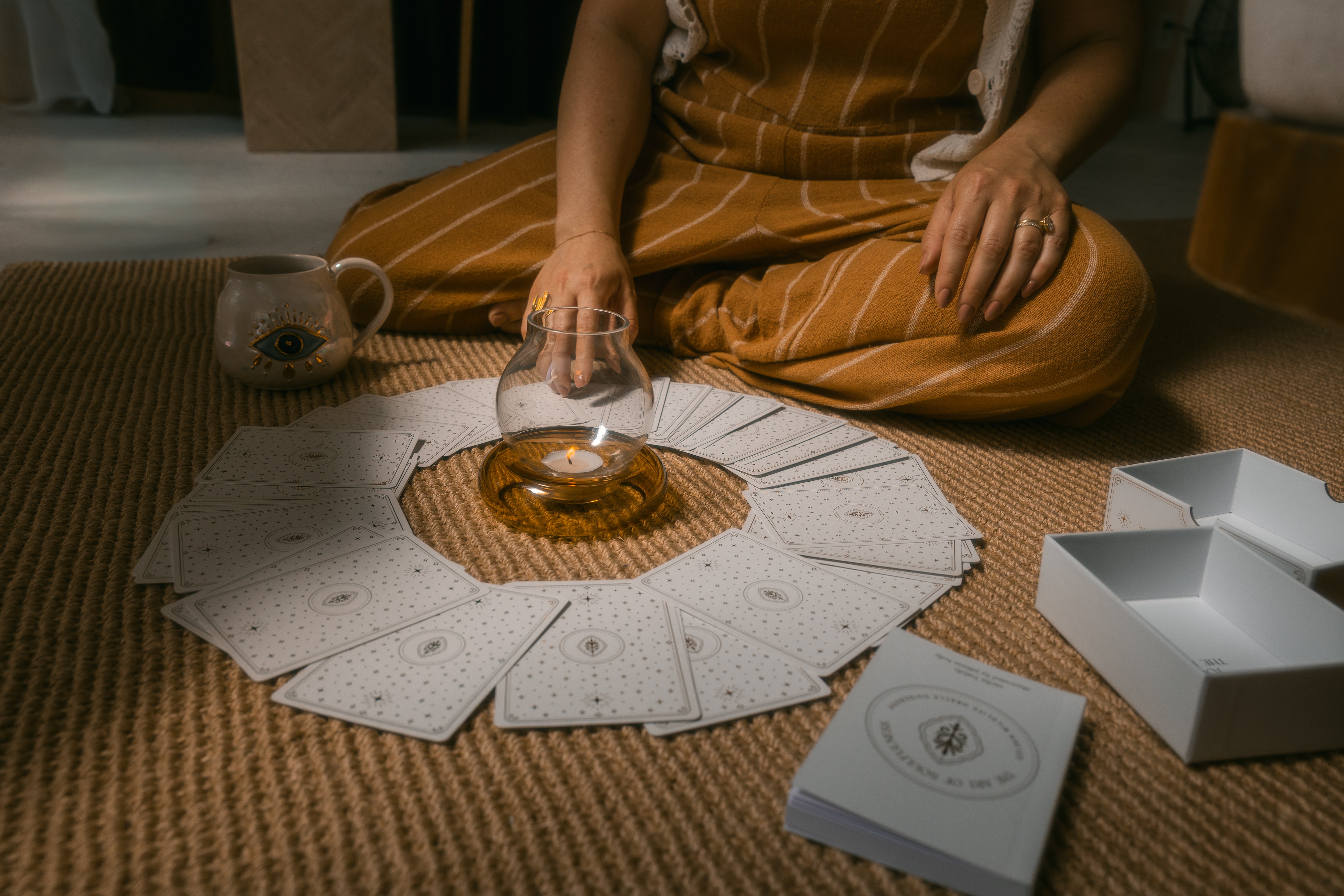 person sitting on a braided carpet reaching to draw a single golden wildlife oracle card from a face-down, circular spread around a candle, with a mug, guidebook and open box nearby.