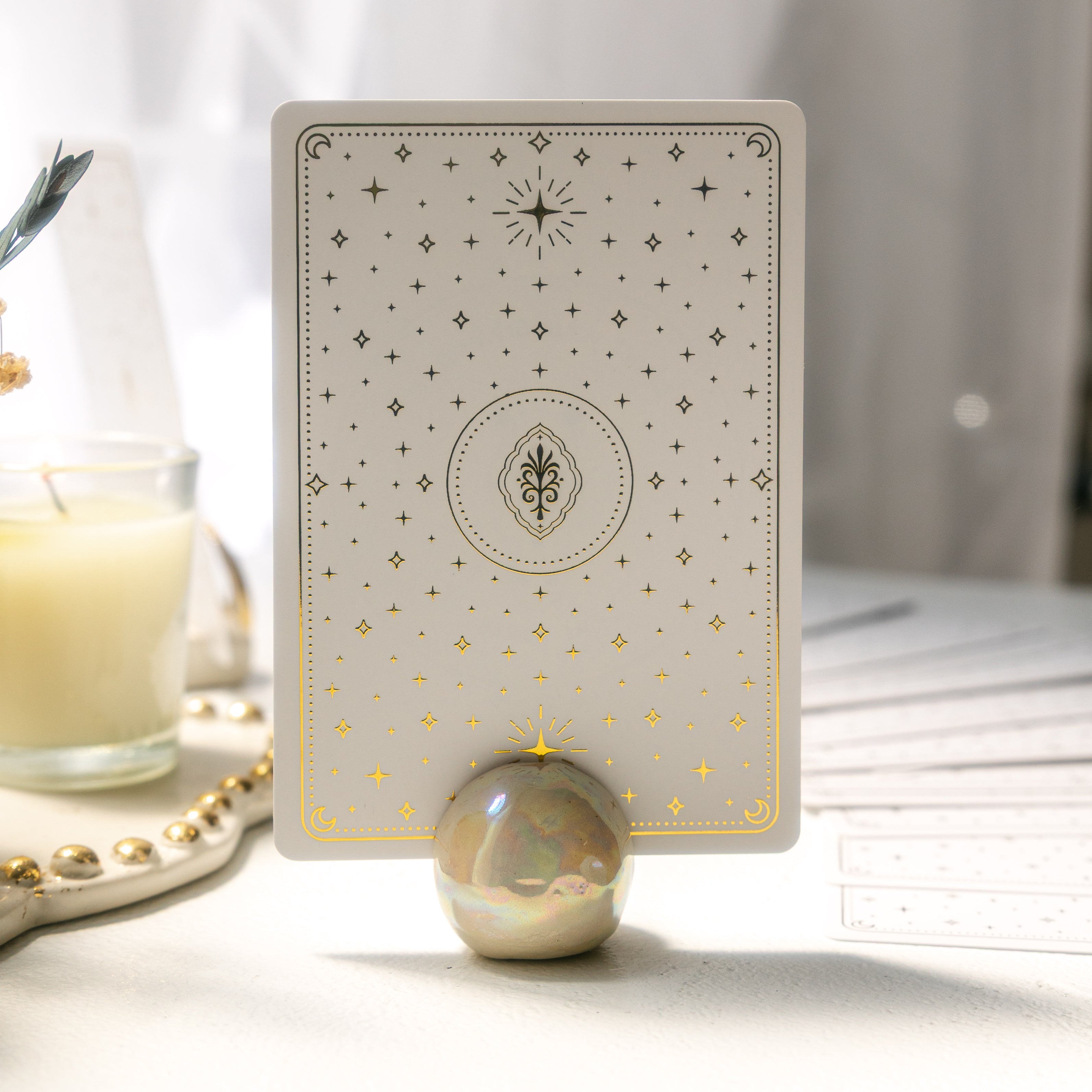 oracle card with a gold foil star design in an iridescent, ceramic orb stand on a white surface near a ritual shelf