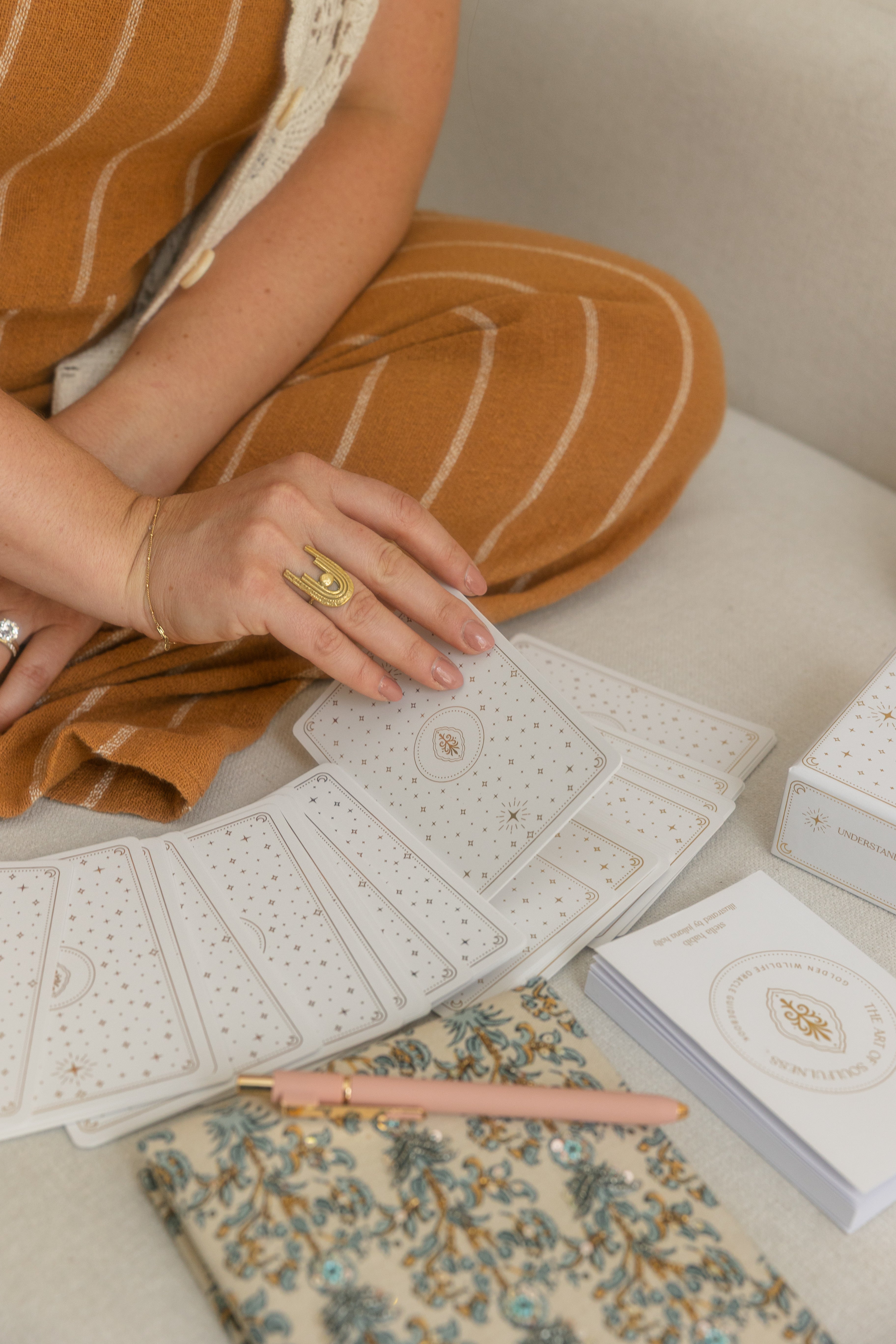 person sitting on a light-coloured couch selecting a golden wildlife oracle card from a face-down arc, wearing a thin gold bracelet, brass ring.