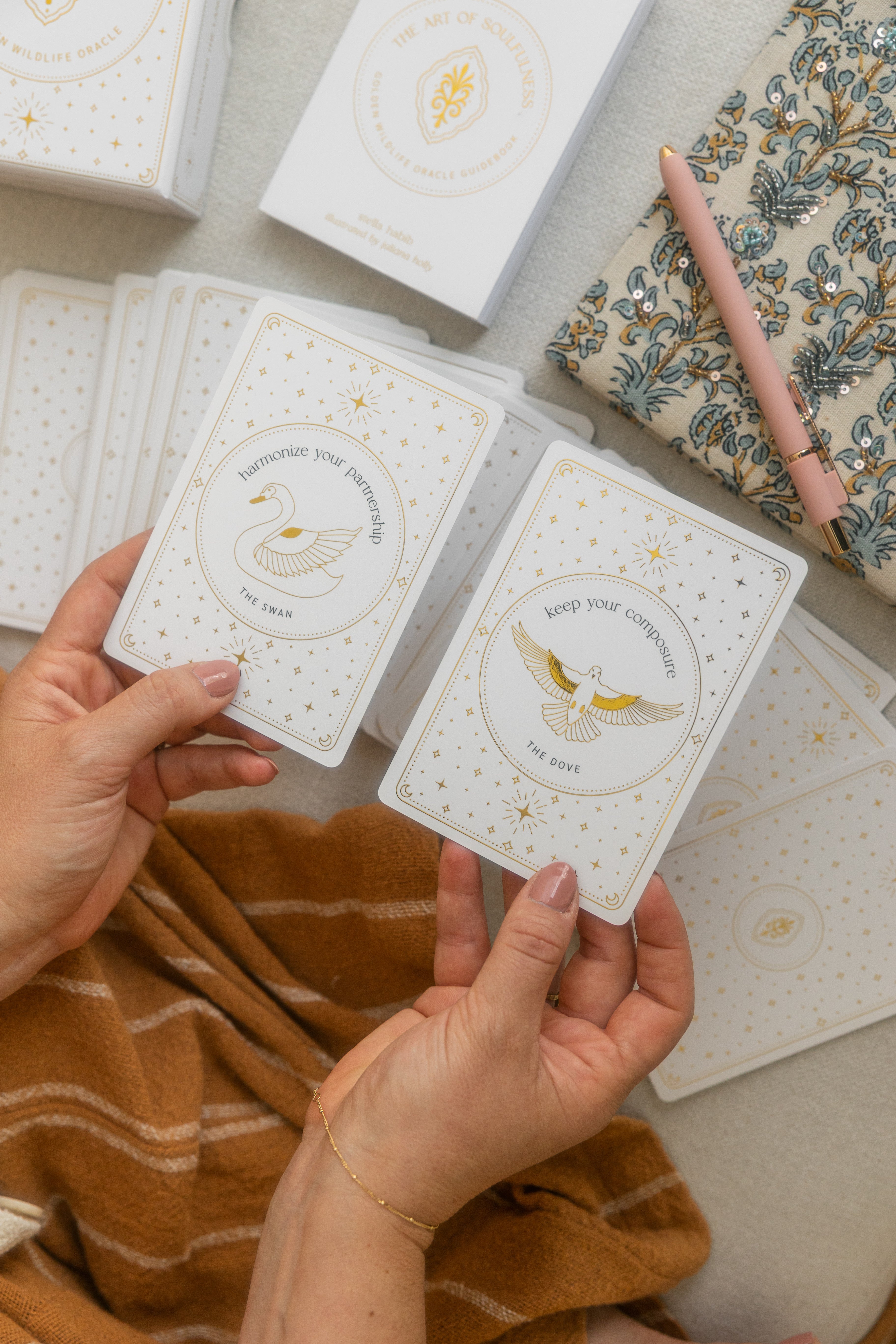 a left and right hand each holding a gold foil oracle card, each with a different message. the right hand card says 'keep your composure' with an illustration of a dove and the left hand card says 'harmonize your partnership' with an illustration of a swan.
