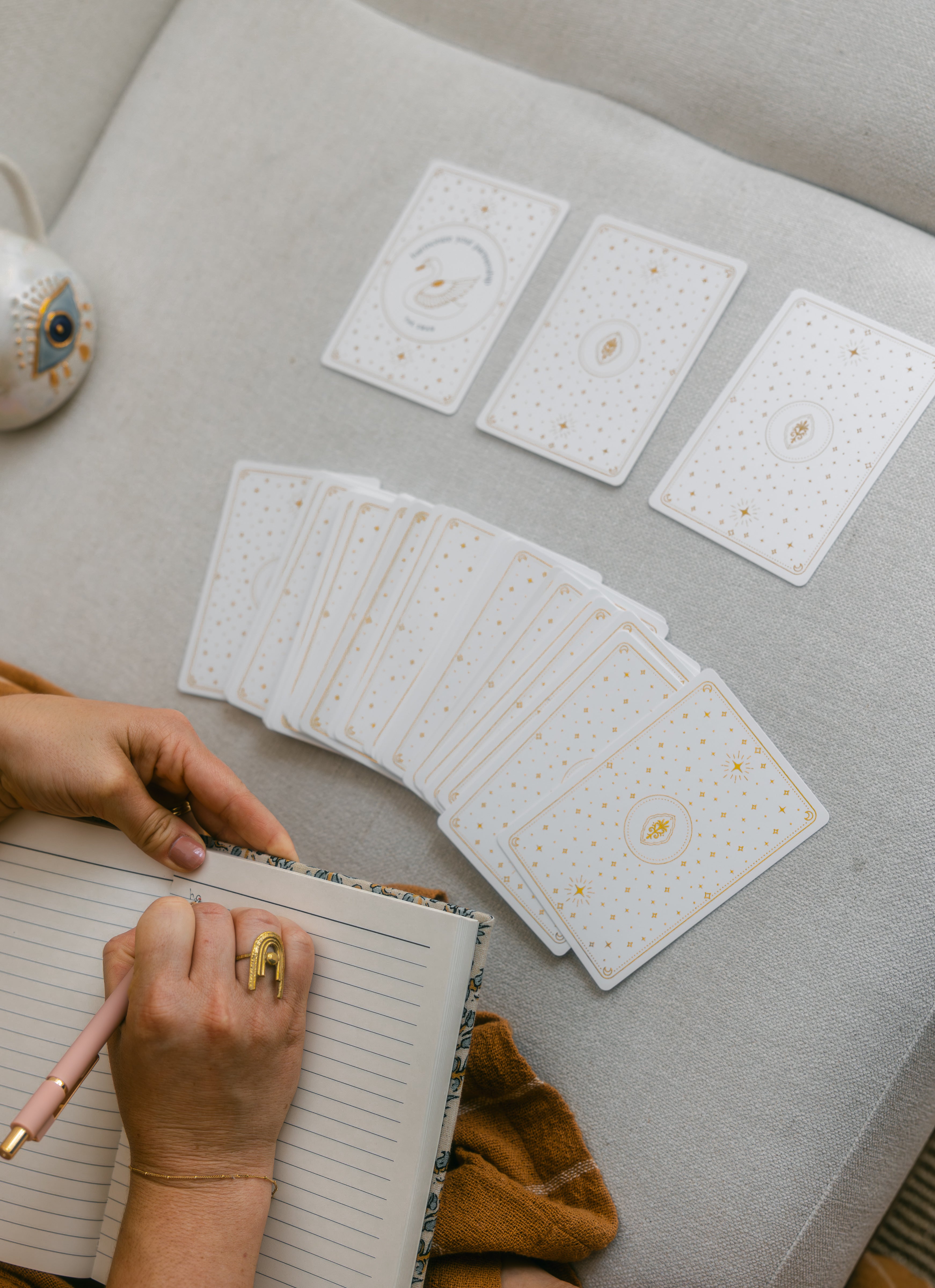 person writing in a notebook with a face-down arc of golden wildlife oracle cards and a three card spread above it, featuring one face-up card with the illustration of a swan.