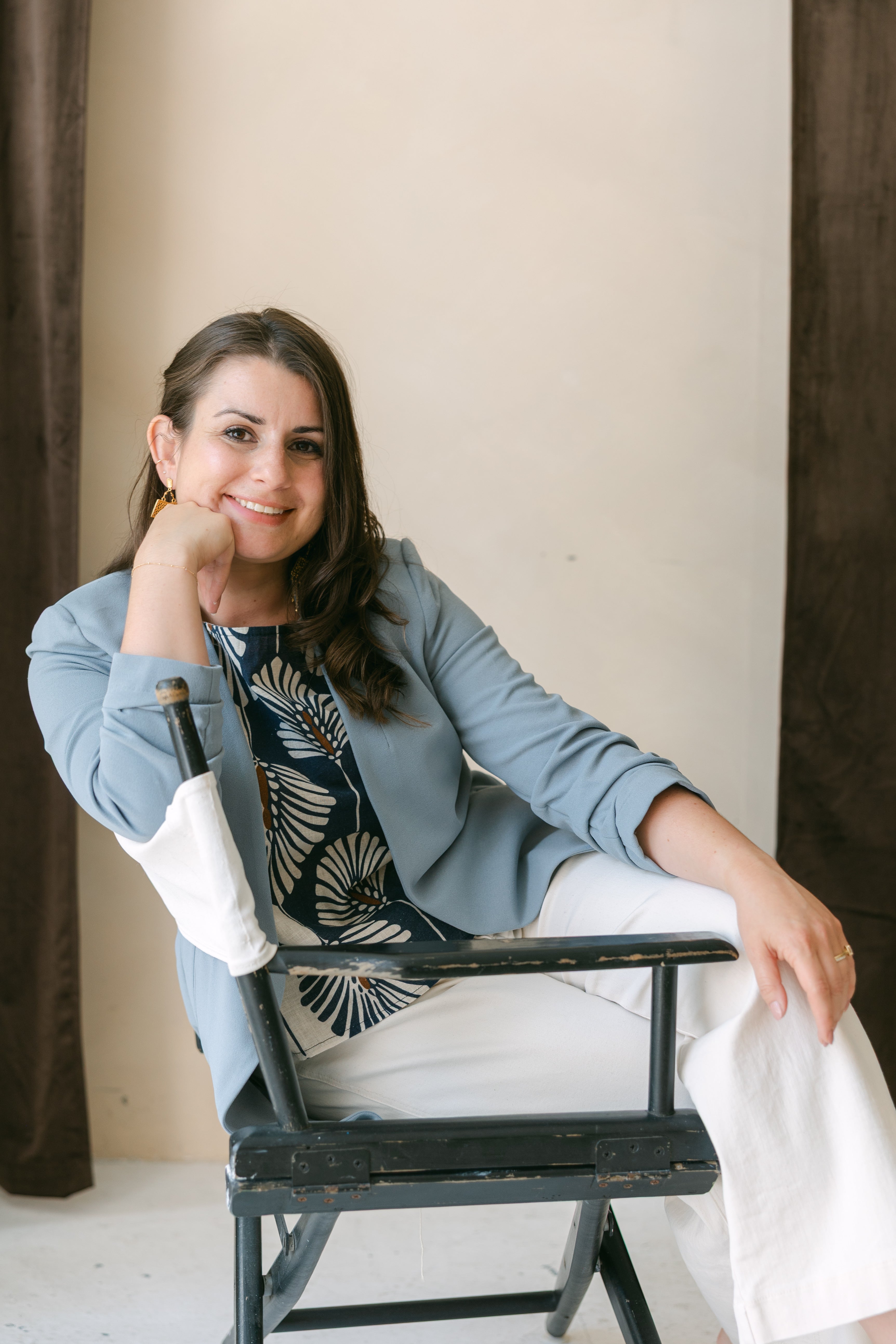 a portrait of stella in a blue blazer, patterned top and white pants, leaning on the backrest of an armchair. 