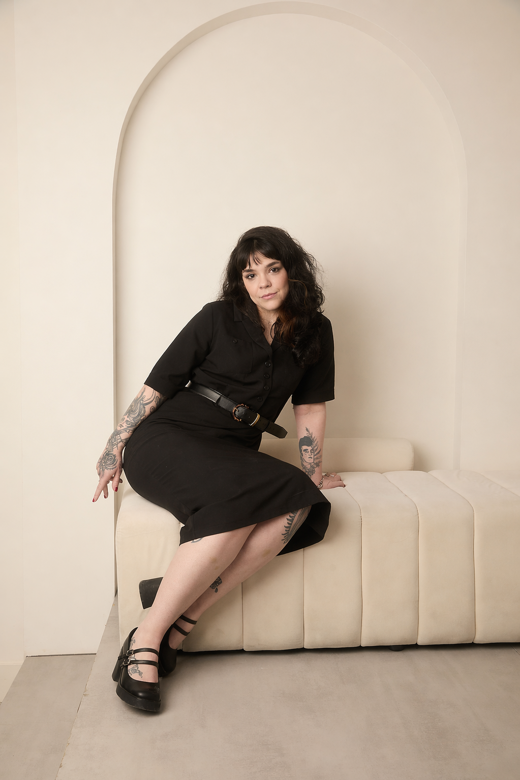 ju wearing a black outfit with black heels, sitting on a modern sofa against a beige wall with arch detailing