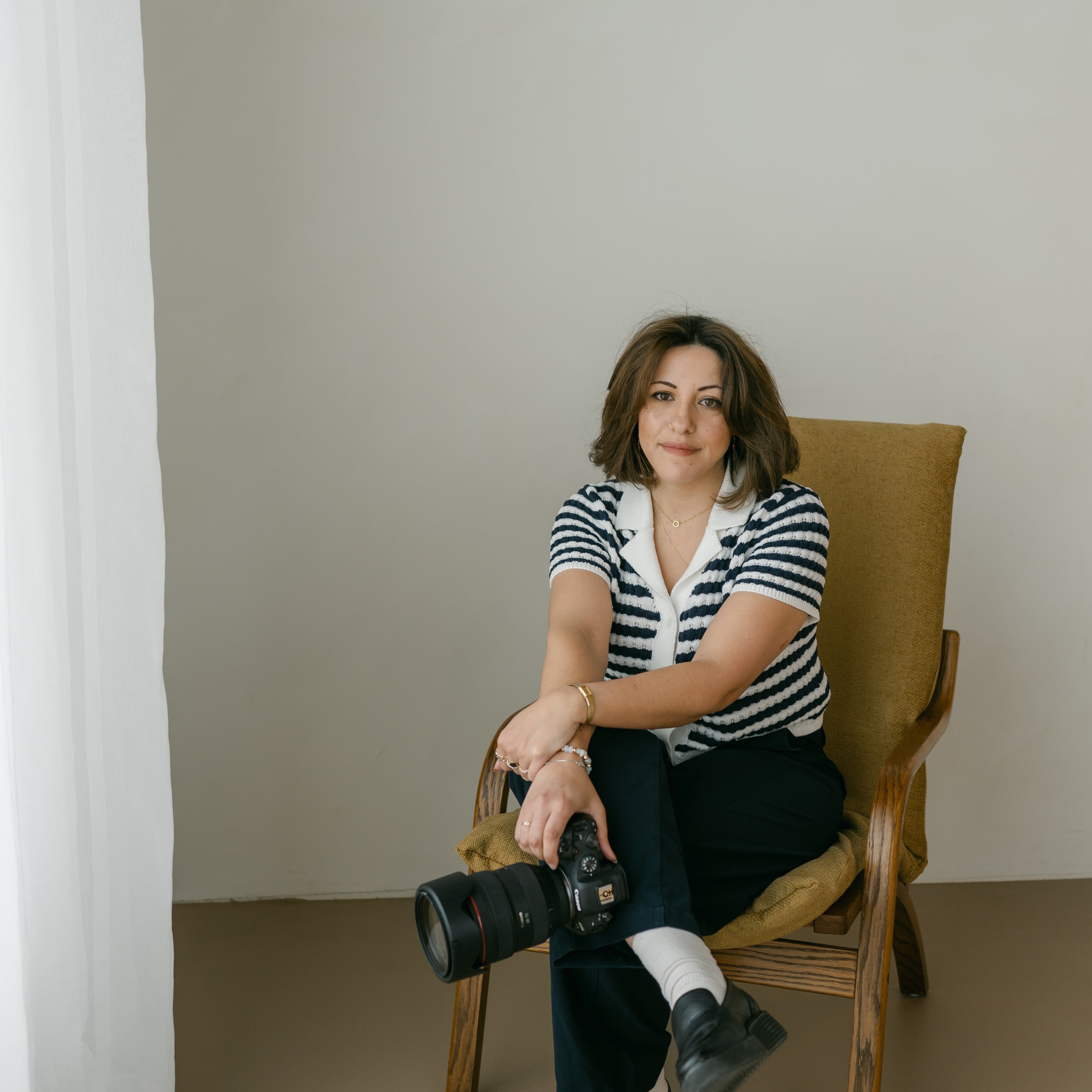 a portrait of kenda sitting in a chair with her legs and wrists crossed, holding a professional camera.