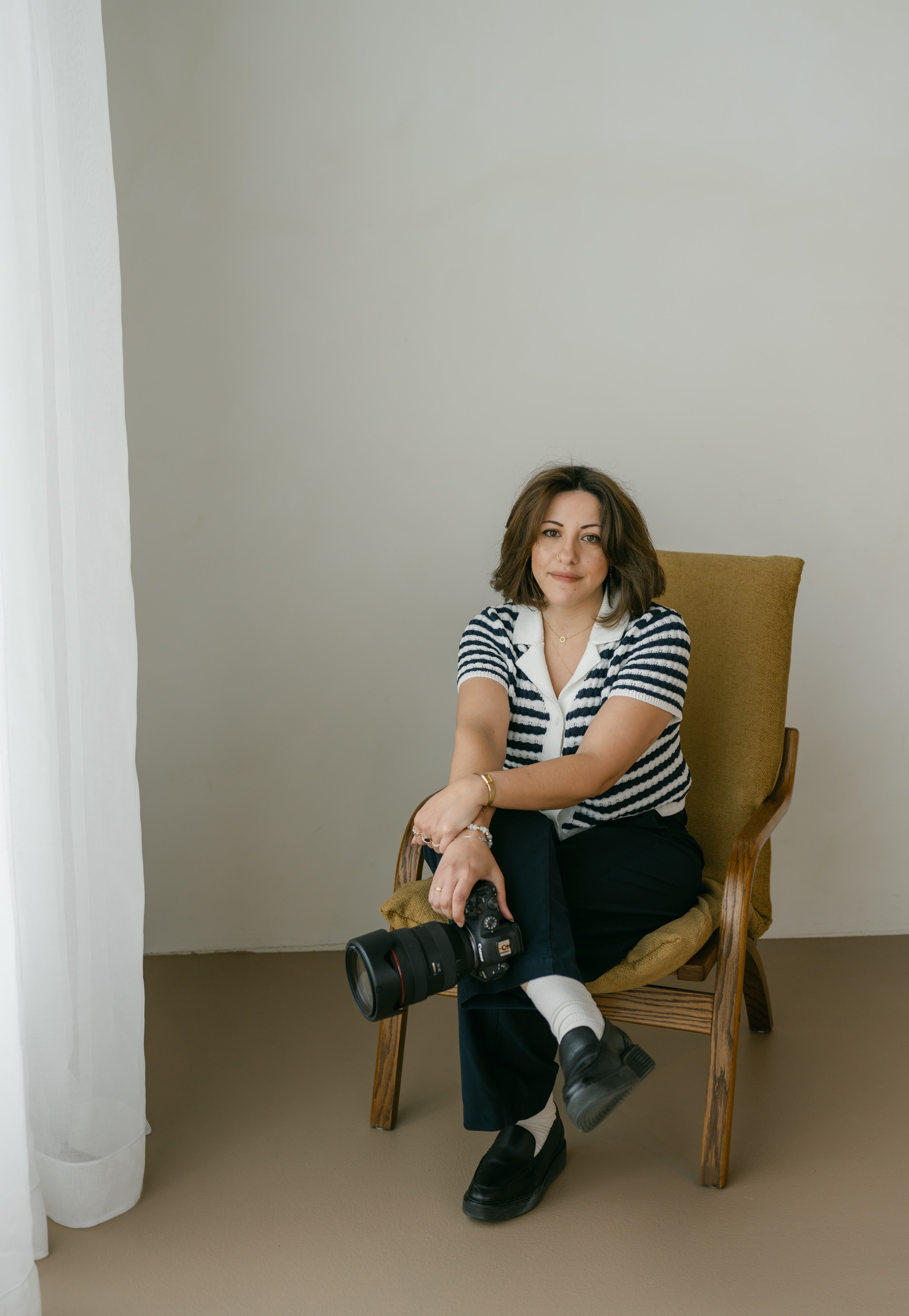 a portrait of kenda sitting in a chair with her legs and wrists crossed, holding a professional camera.