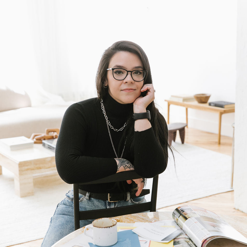 a portrait of ju leaning against the backrest of a chair near a table with a coffee mug, stack of books and magazine clippings.