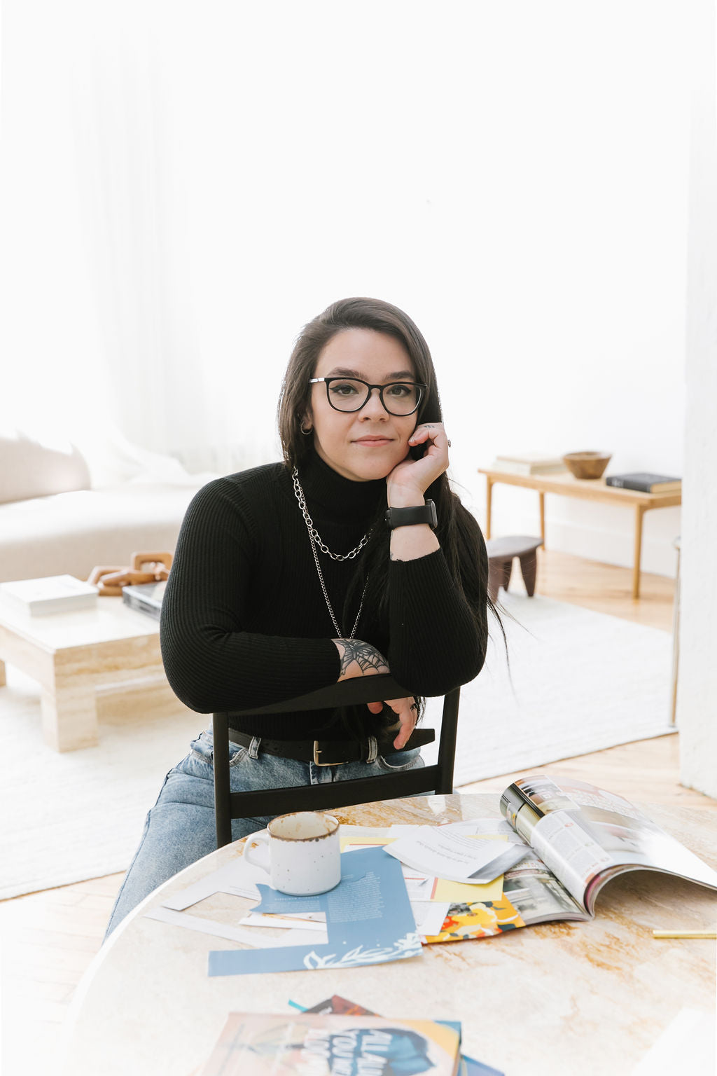 a portrait of ju leaning against the backrest of a chair near a table with a coffee mug, stack of books and magazine clippings.